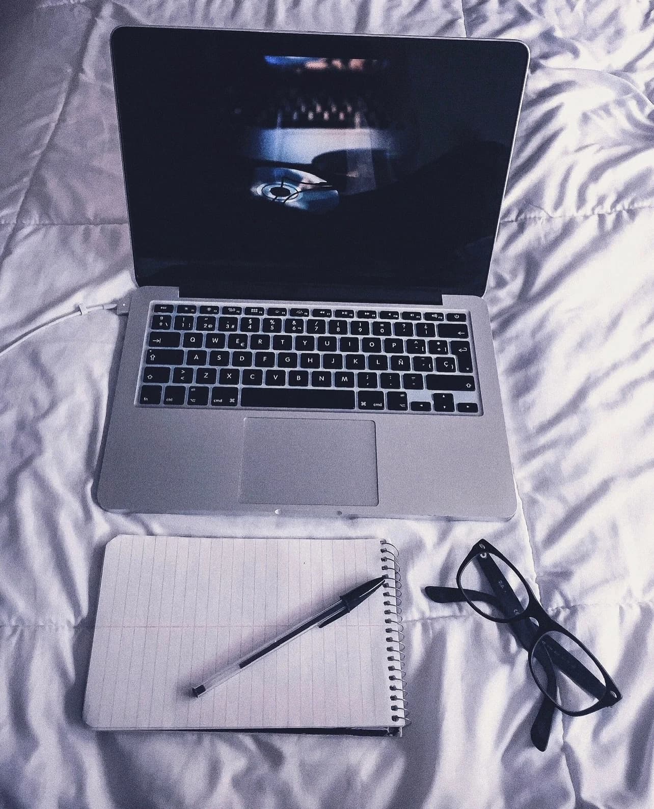 Laptop, notebook and glasses arranged on white bedding