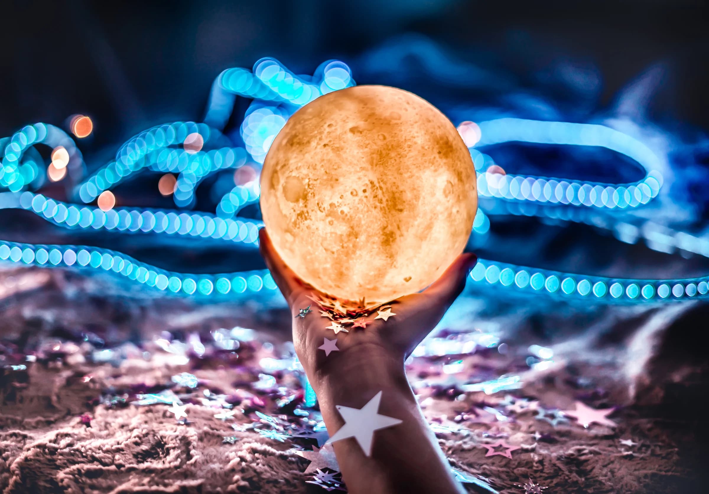A glowing moon held in a hand against blue string lights